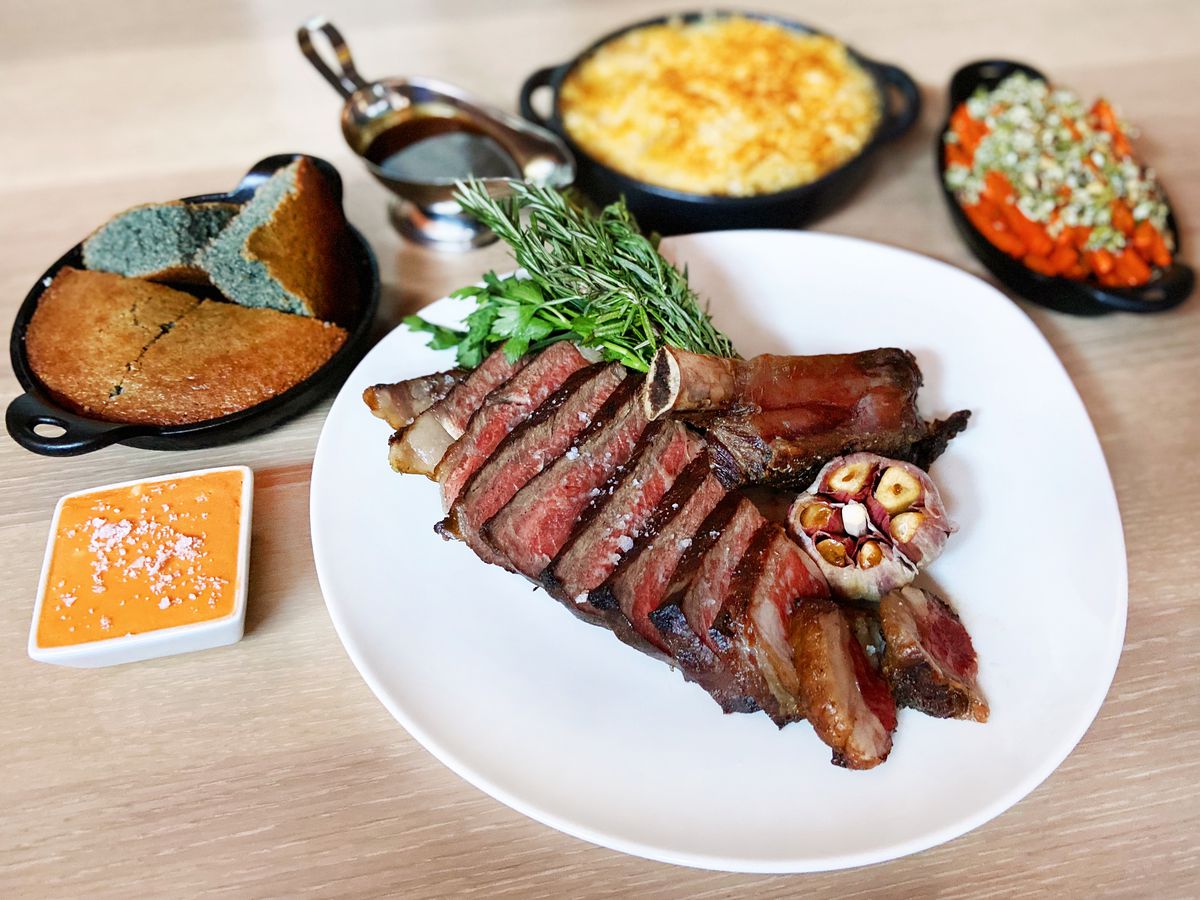 A rib-eye steak sliced with a clove of roasted garlic and thyme garnish, beside cornbread, mac and cheese, and other sides