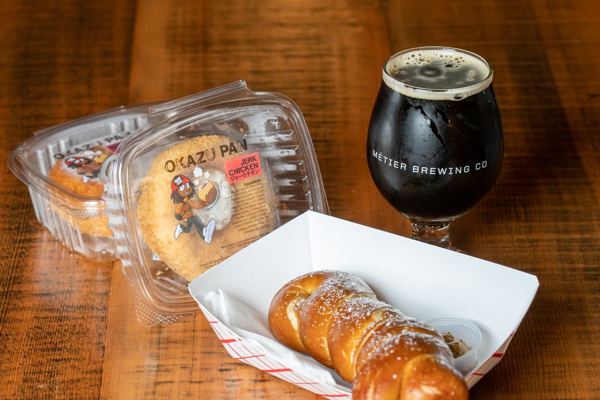 A pretzel twist in a paper boat and two okazu pan (fried bread rolls) in plastic containers next to a glass of jet-black beer on a wooden table.