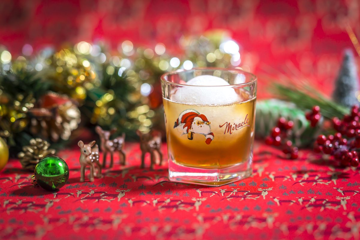 A glass that says Miracle and has a Santa graphic has a snowball and brown liquor in it. It’s displayed on a Christmas-themed backdrop.