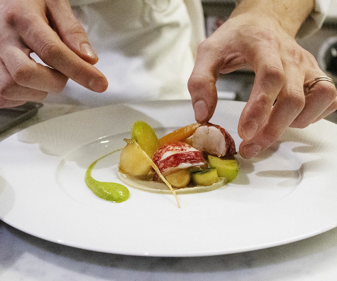 A chef places thick cuts of lobster in a bed of other ingredients on a mostly clean white plate