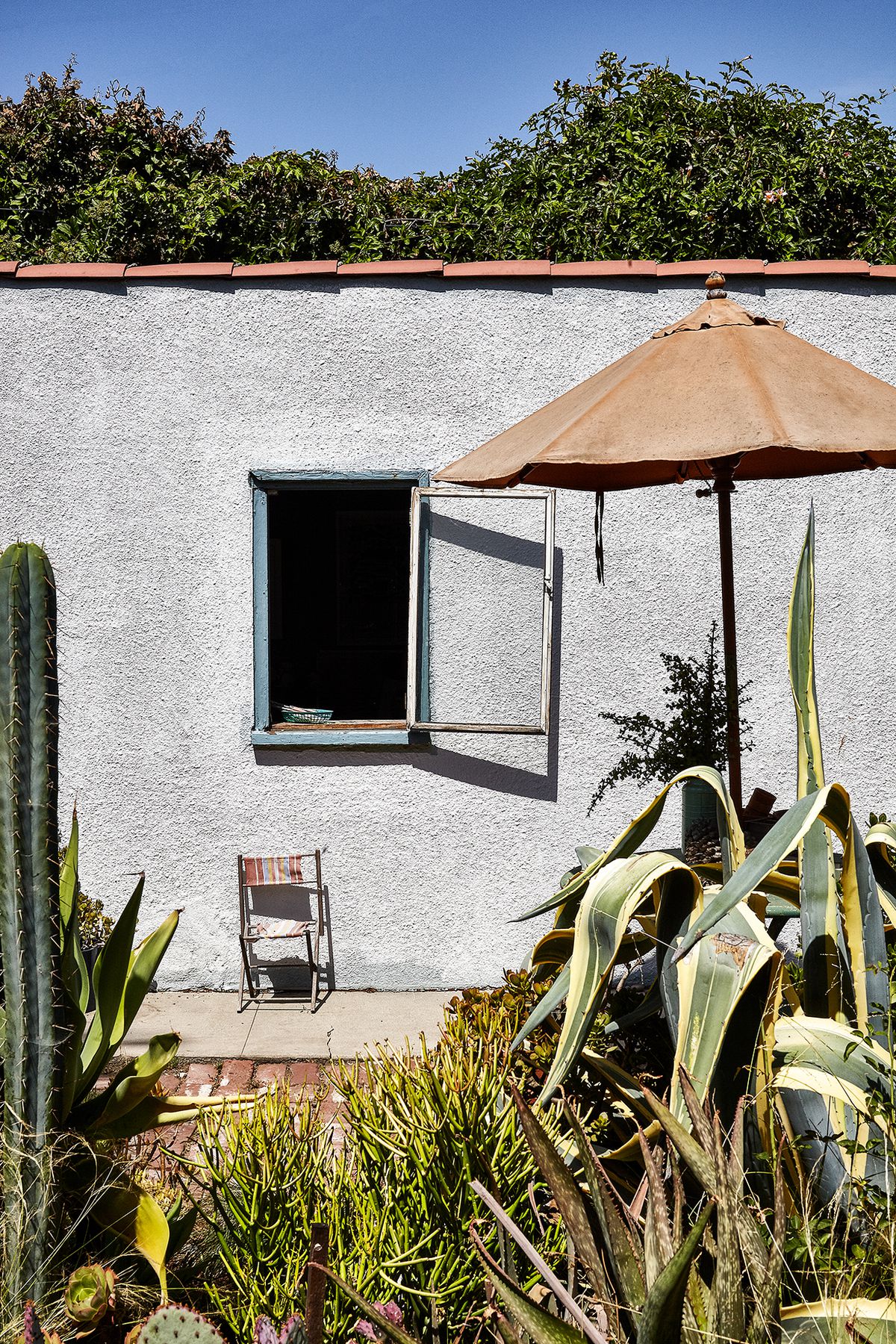 Detail of the side of a studio attached to a home in LA. The building is grey with a red tile roof. There is one window, with blue trim and it is open, with a little chair outside of it on the ground. An umbrella on top of a table and lots of cactus and plants in the foreground. 