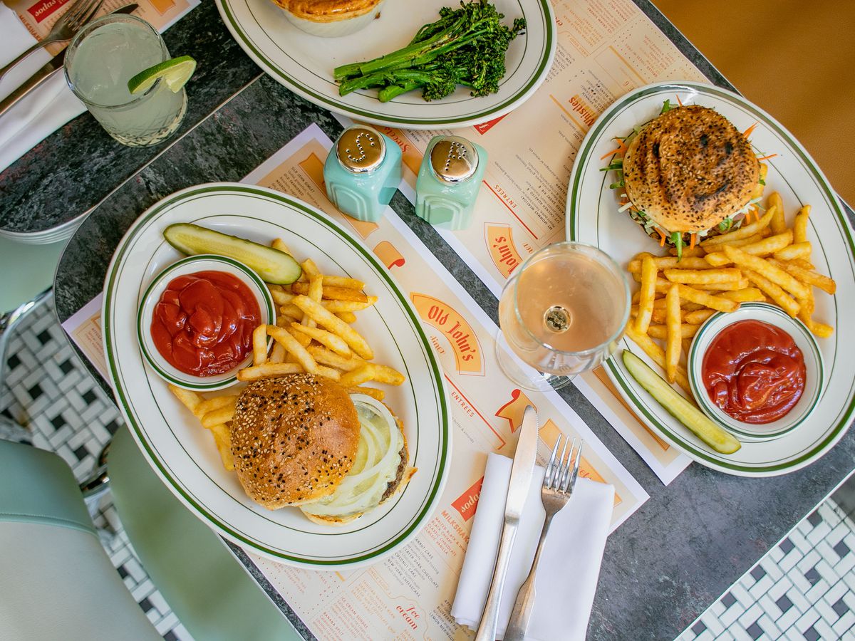 An overhead shot of oval dishes with burgers, french fries, pickles, and greens laid out across two blue tables with booths and chairs on the side
