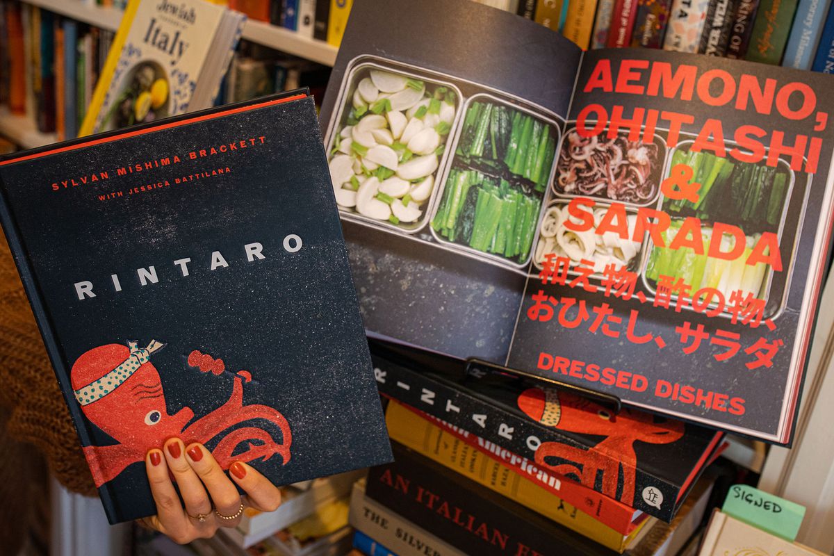 A copy of the cookbook “Rintaro: Food and Stories from a Japanese Izakaya in California” is opened to a page displaying food from Rintaro, while a hand holds a closed copy of the book next to the opened pages.