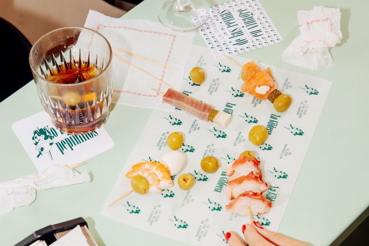 People pick at skewered snacks on a branded placemat while drinking glasses of sherry.