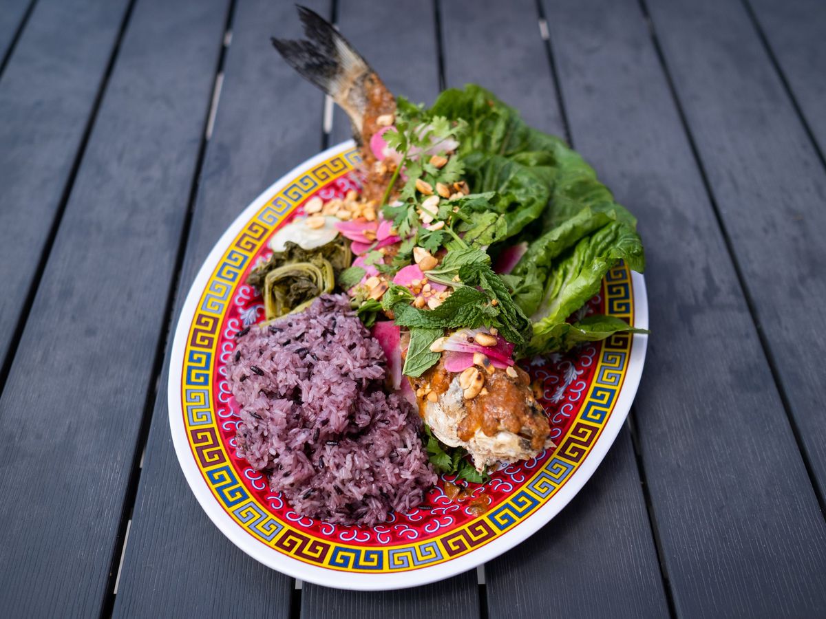 Grilled whole fish with purple rice and greens on a red plate on a grey wooden table. 