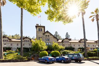Das Gebäude des Culinary Institute of America, das einer Burg mit Turm ähnelt. Im Vordergrund sind Bäume und ein Parkplatz zu sehen. Die Sonne scheint hell durch die Bäume.
