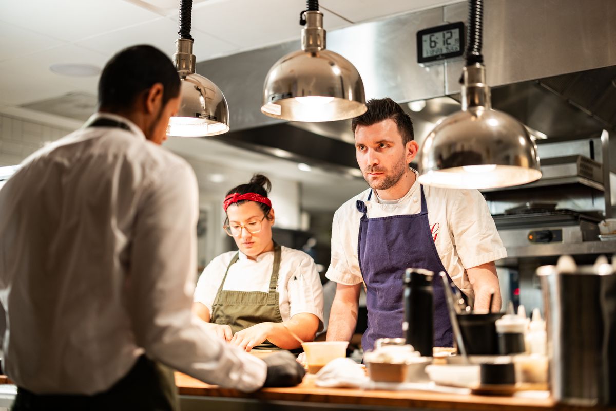 Two chefs stand behind a counter. One, a woman on the left, is looking down and working. The other, a man on the right, is looking intently at a man in an apron and white shirt across the counter.