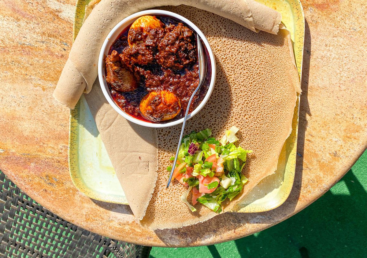 Bird’s-eye view: A dish of doro wat, slow-cooked chicken with onions and spices, sits atop a larger plate next to injera and salad. The background is a beige-colored tabletop.