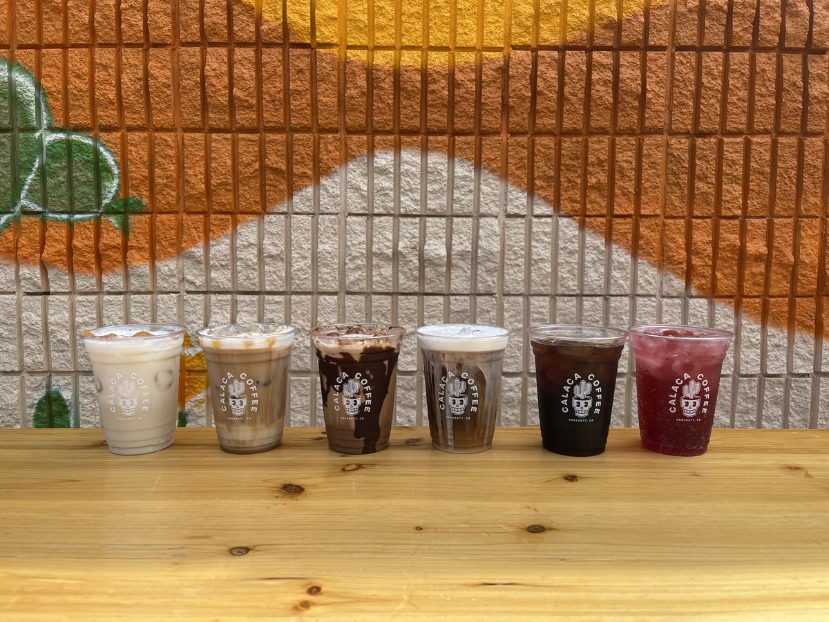 Six different coffee drinks from Calaca Coffee are lined up on a table in front of a colorful mural and background.