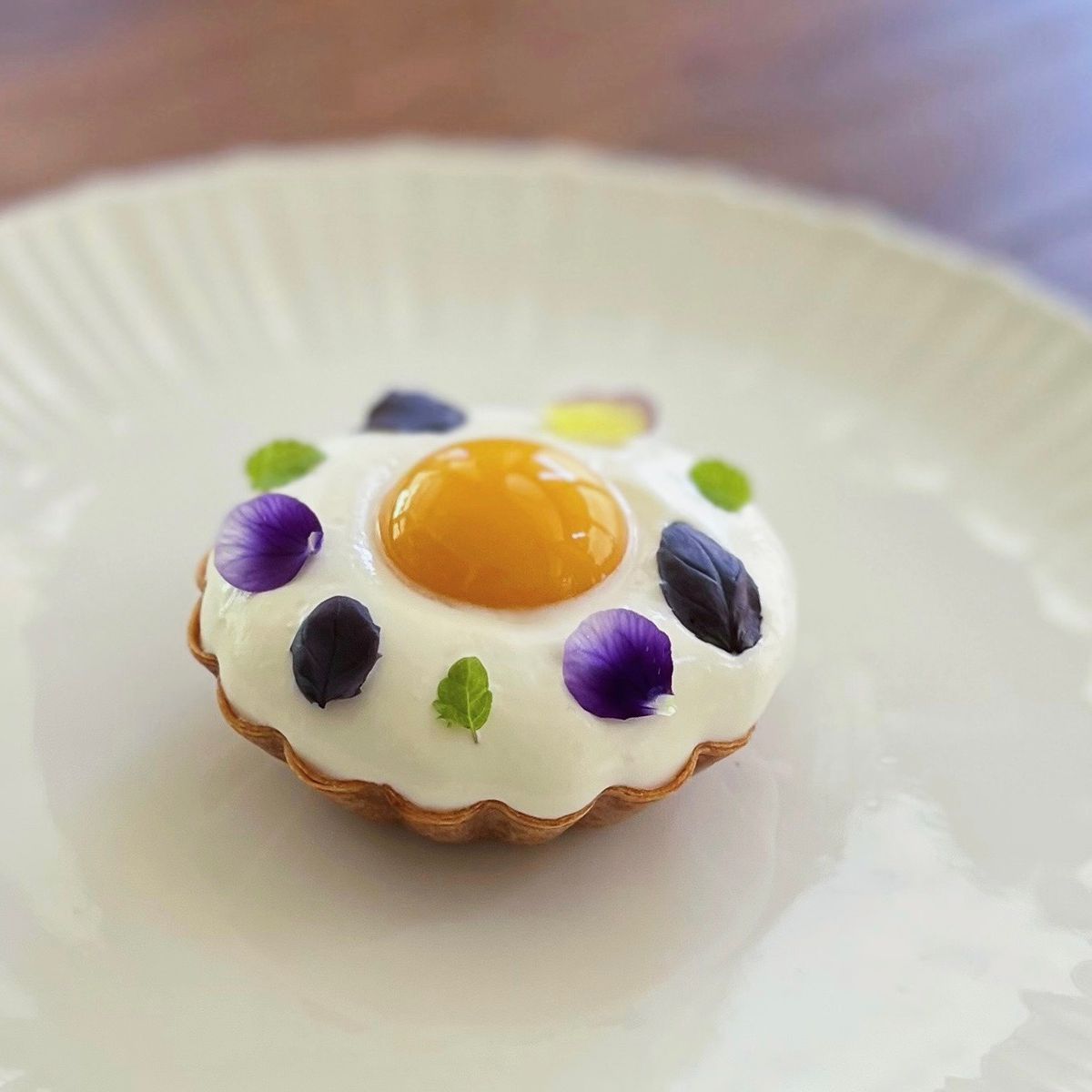 A plate holds a round “egg tart” made to look like an over easy egg, with petals of flowers atop the egg white portions.