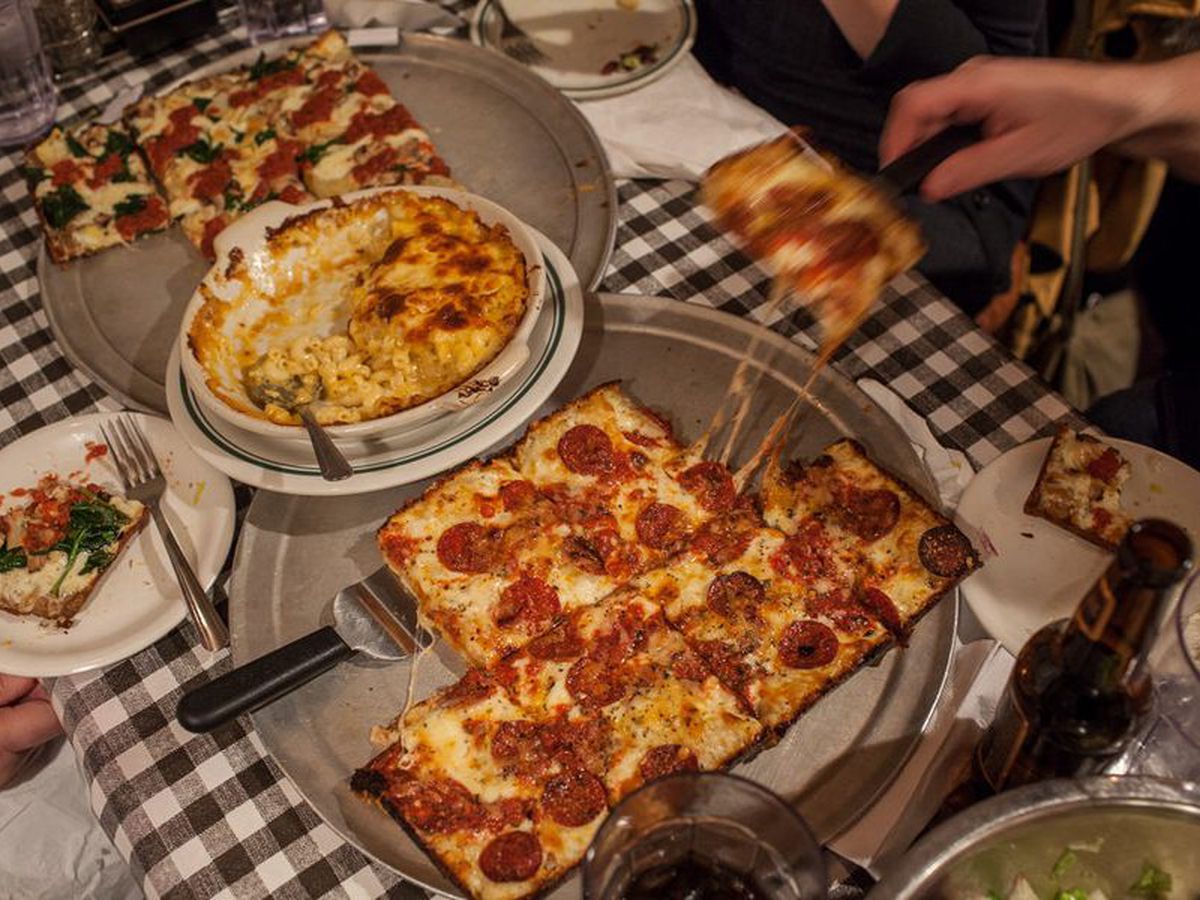 a square pepperoni pizza being served onto plates over a black and white checked tablecloth with macaroni and cheese in a dish.