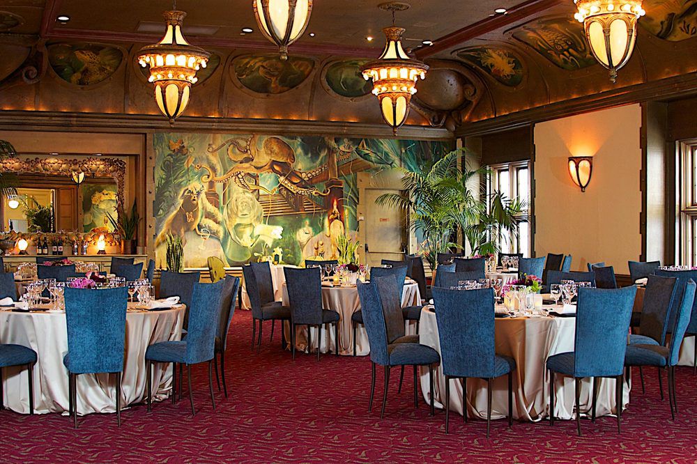 A ballroom decorated with chandeliers, a mural with animals, and round tables fit for large groups.