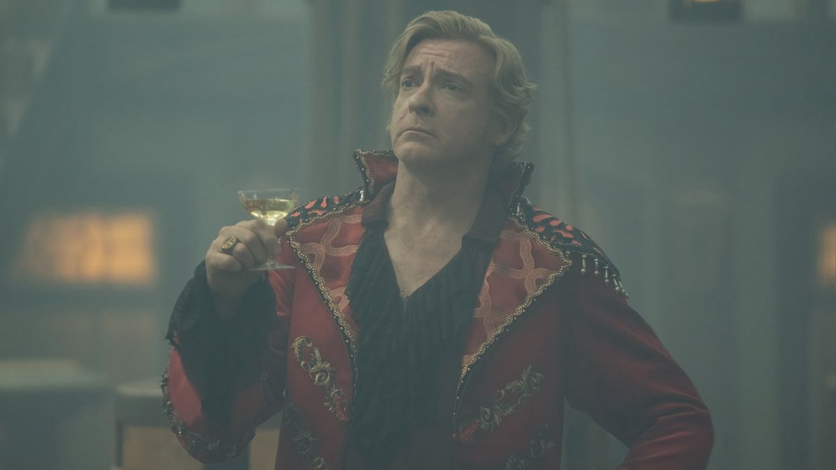 Stede Bonnet (Rhys Darby) poses with a cocktail, looking smug in a tacky, rickrack covered red silk jacket in season 2 of Our Flag Means Death