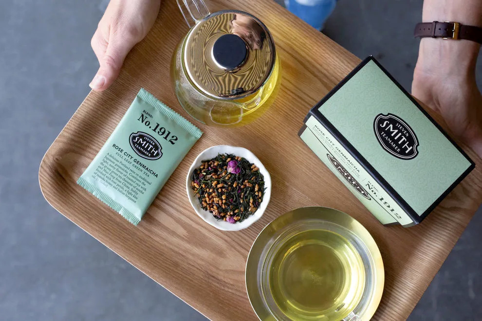 Hands carry a tray with a pot of tea and a box of Rose City Genmaicha at Smith Teamaker.
