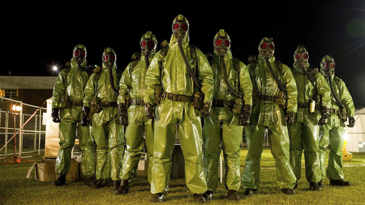 A group of ominous figures in lime-green hazmat suite and gas masks with red tinted lenses.