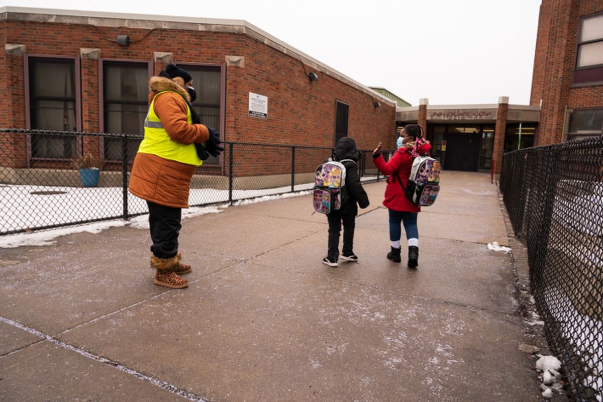 A woman, wearing a long orange winter coat and a crossing guard vest, greets two young students entering the school.