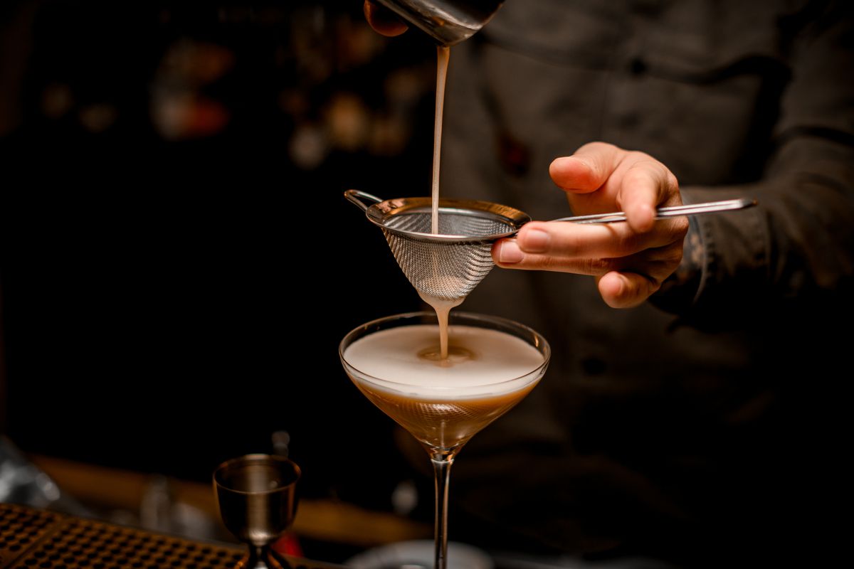 The hands and torso of a bartender in a black chef’s coat, using a metal sieve to strain an espresso martini into a martini glass on a bar top.