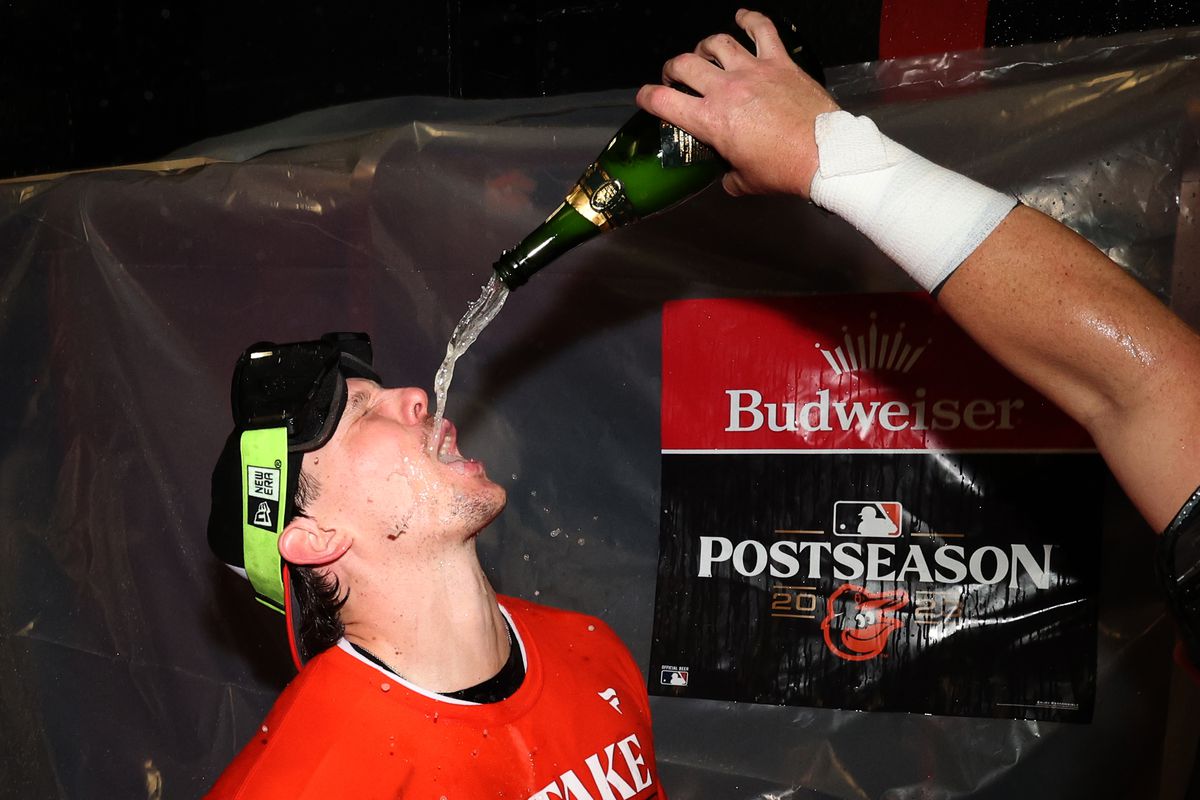 Orioles player Adley Rutschman, wearing an orange “Take October” shirt, stands with head tilted back and mouth open as beer is poured from a bottle into his mouth. He has goggles on his forehead that are not shielding his eyes.