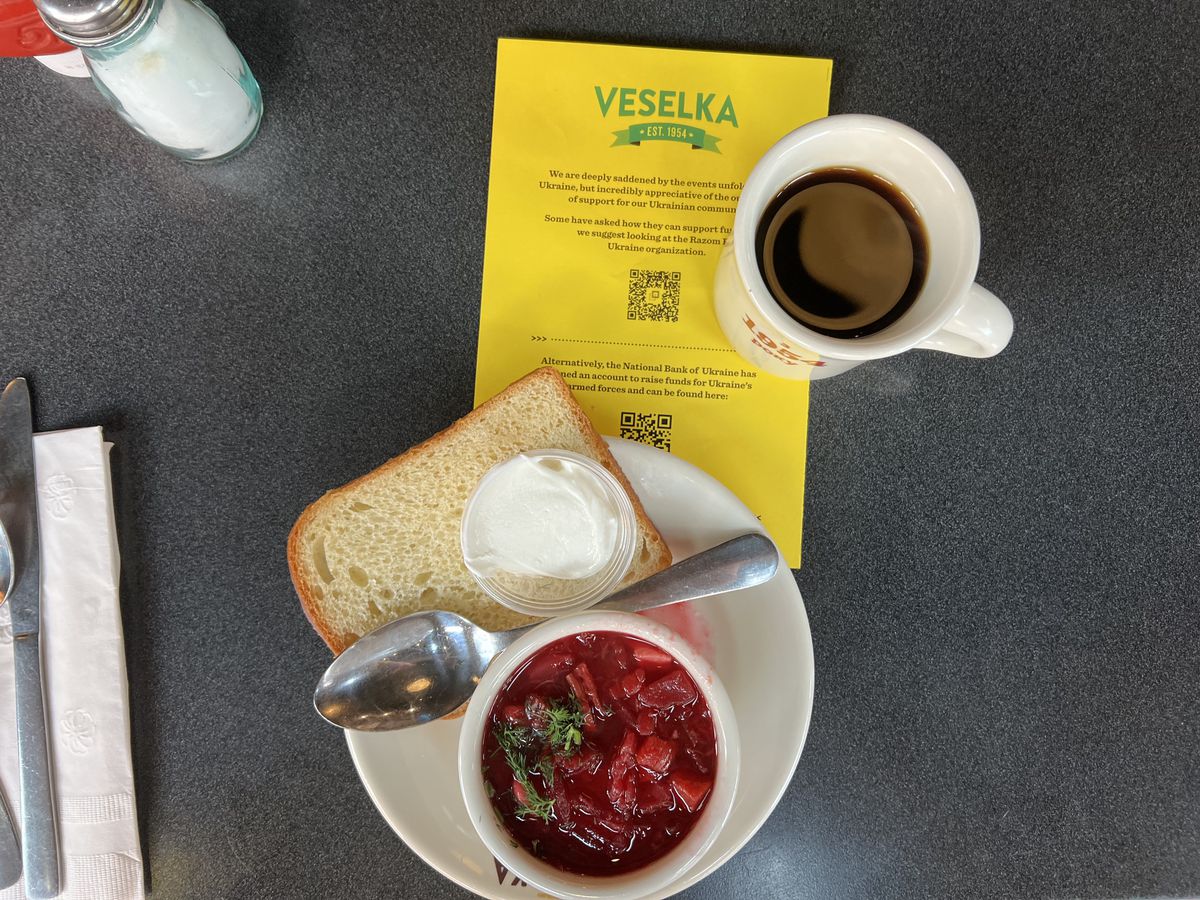 A cup of bright red borscht on a white plate with a  slice of bread and sour cream and spoon next to a yellow flyer and cup of coffee.