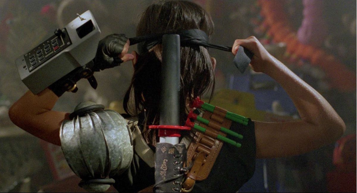 A young boy, viewed from behind, ties a bandana around his long hair. He is loaded up with toy weapons in Deadly Games.