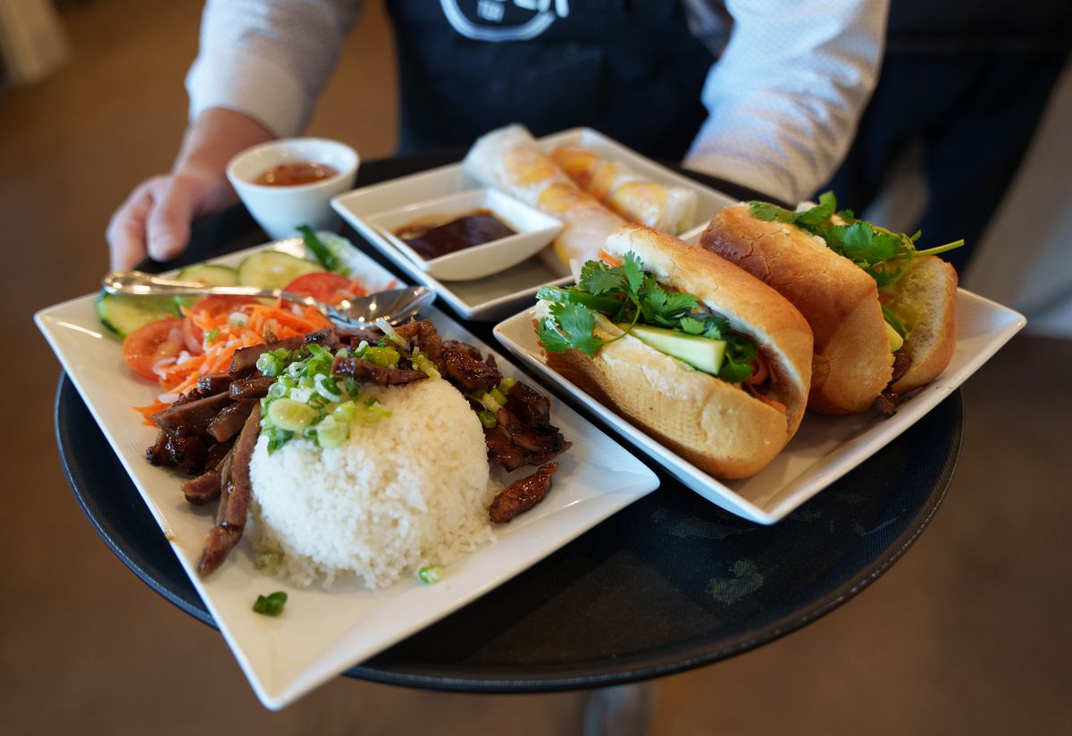A person carries a tray with a pork and rice dish, banh mi, and spring rolls.