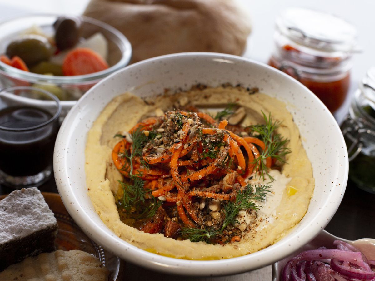 A bowl spread with hummus and topped with shaved carrots, spices, herbs, spots of olive oil, and a small dollop of tahini on a table with other blurred items gathered around like pickles and a glass of wine