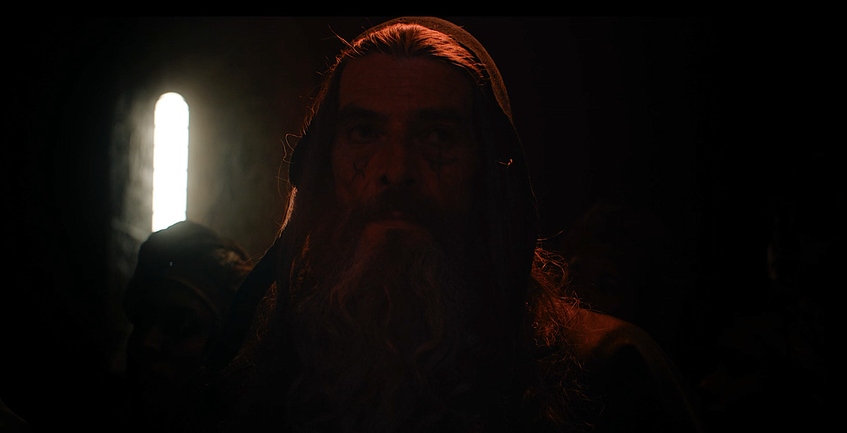 Merlin, in dim, rich red light, casts a spell in David Lowery’s The Green Knight