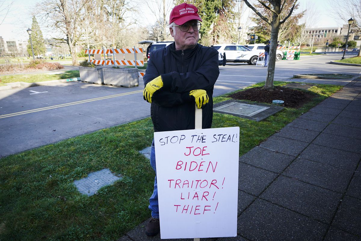 A person wearing a MAGA hat stands on a sidewalk and leans on a sign that reads, “Stop the steal. Joe Biden: Traitor! Liar! Thief!”