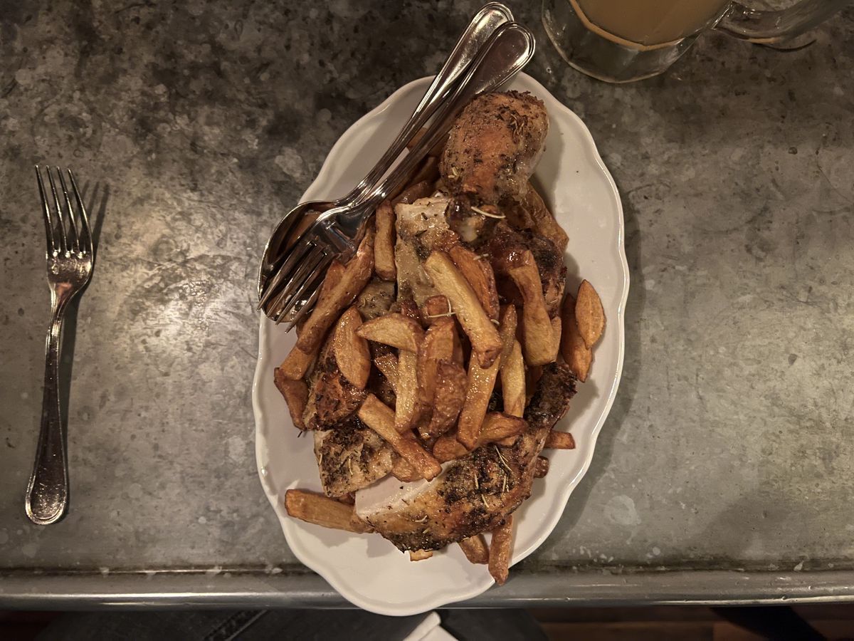 Fries sit above cut roast chicken on a white plate on a silver bar counter; a fork and a spoon sit on the plate