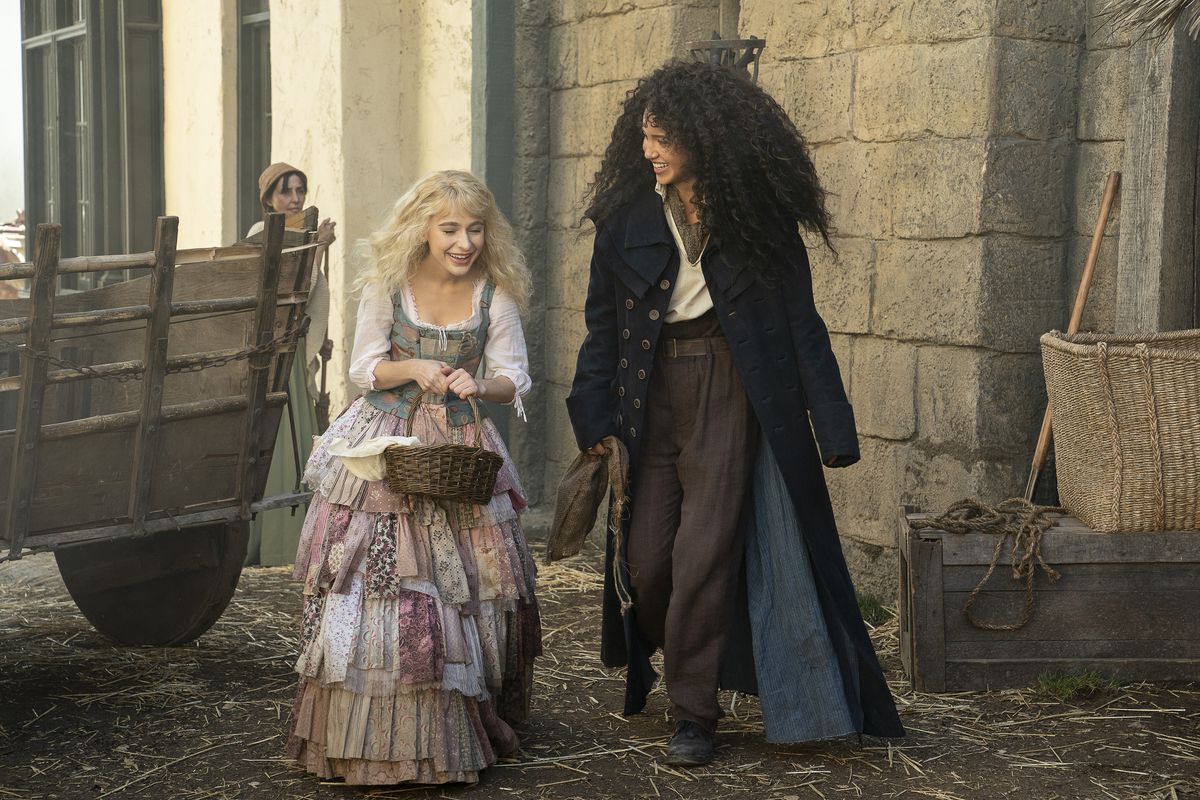 a blonde girl in a dress walking with a taller girl with curly hair who wears a long coat; they’re walking through a fantasy village