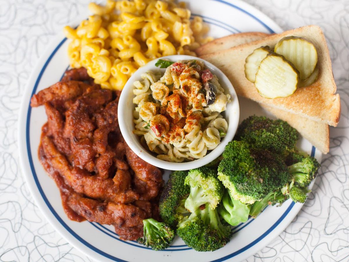 A white plate holds a seasoned chicken, mac and cheese, broccoli, bread with pickles, and in the middle a bowl of pasta salad.