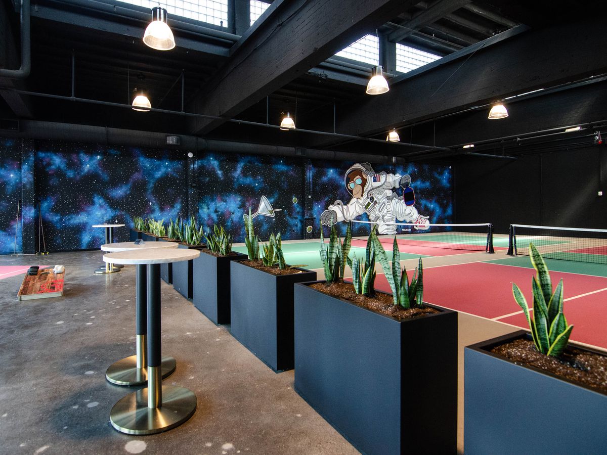 An indoor pickleball court with cornhole and some high-top tables. A mural in the background features a chimpanzee in an astronaut suit floating through space.
