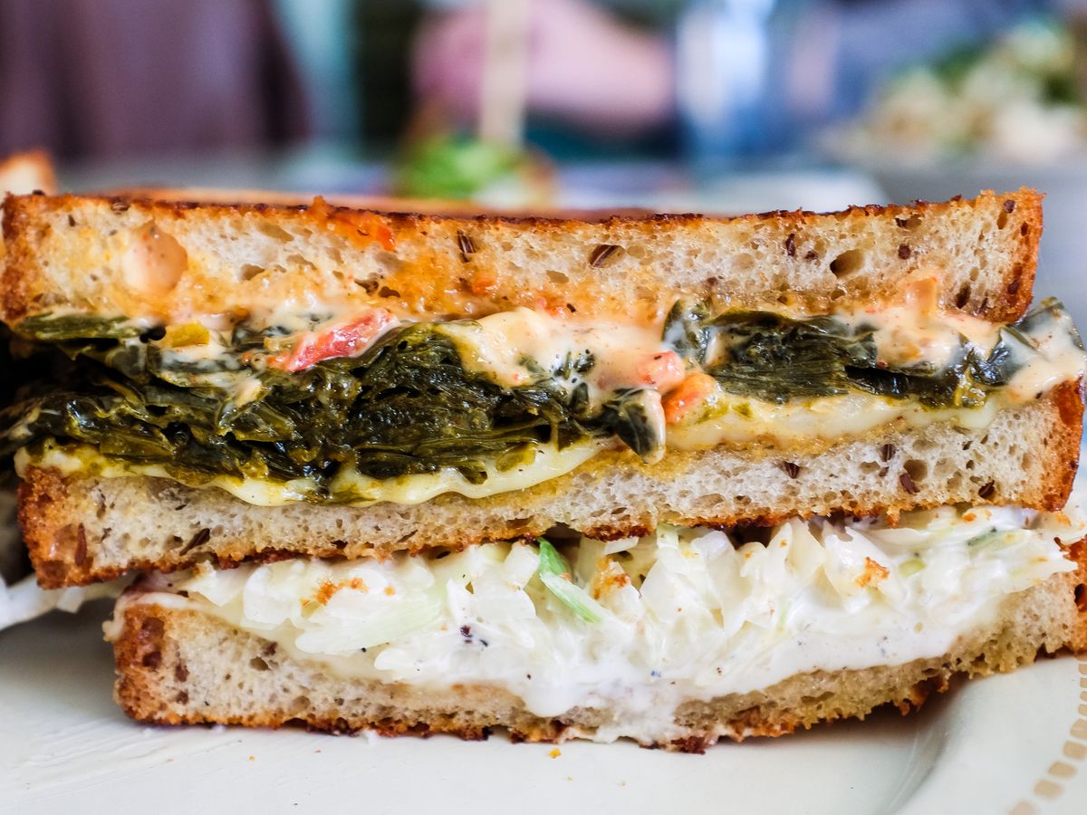 A sandwich of Rye bread, coleslaw, collard greens, and sauce is cut in half on a white plate. 