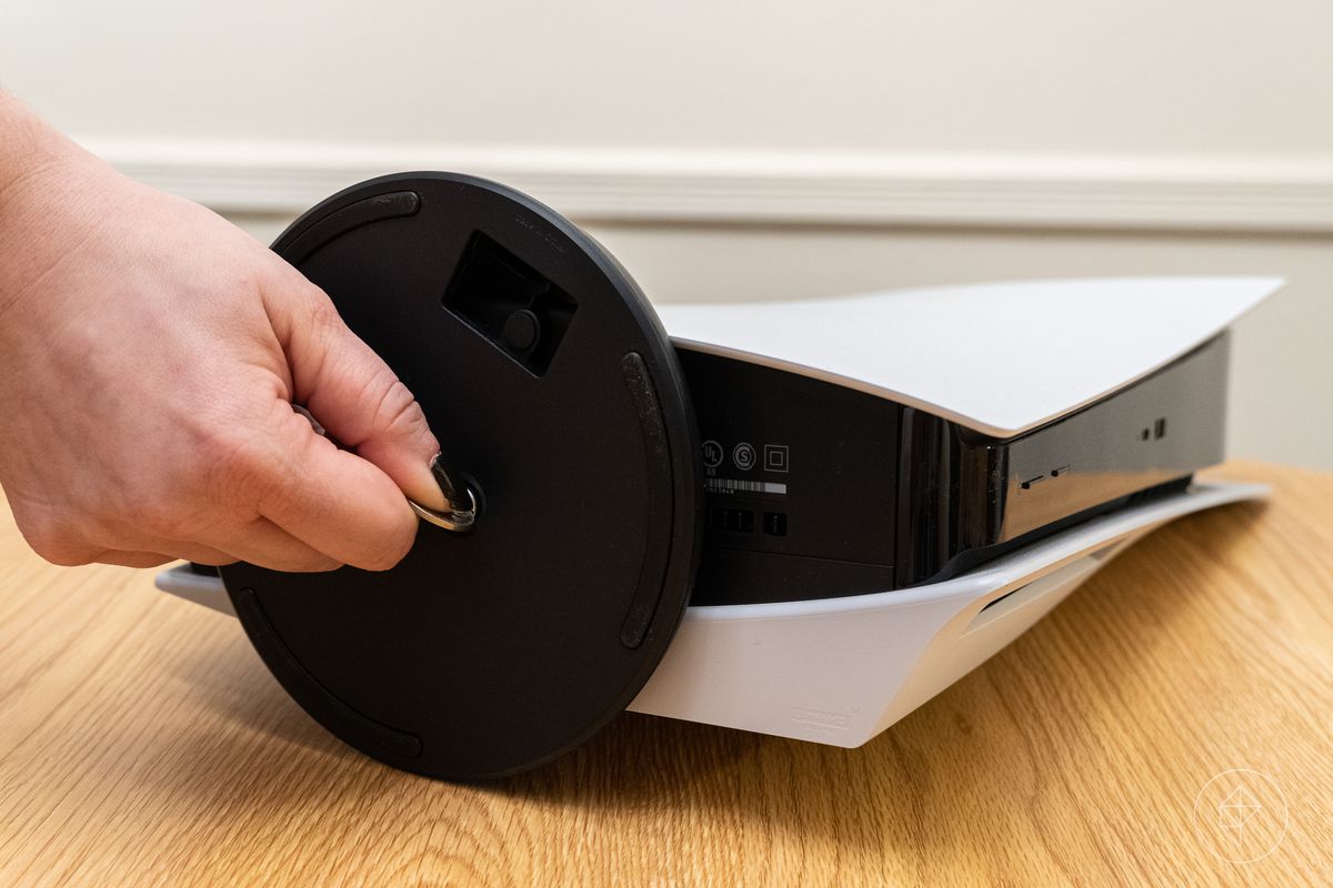 a hand holds a coin to turn the screw of the PlayStation 5 stand, with the console lying horizontally on a wooden table