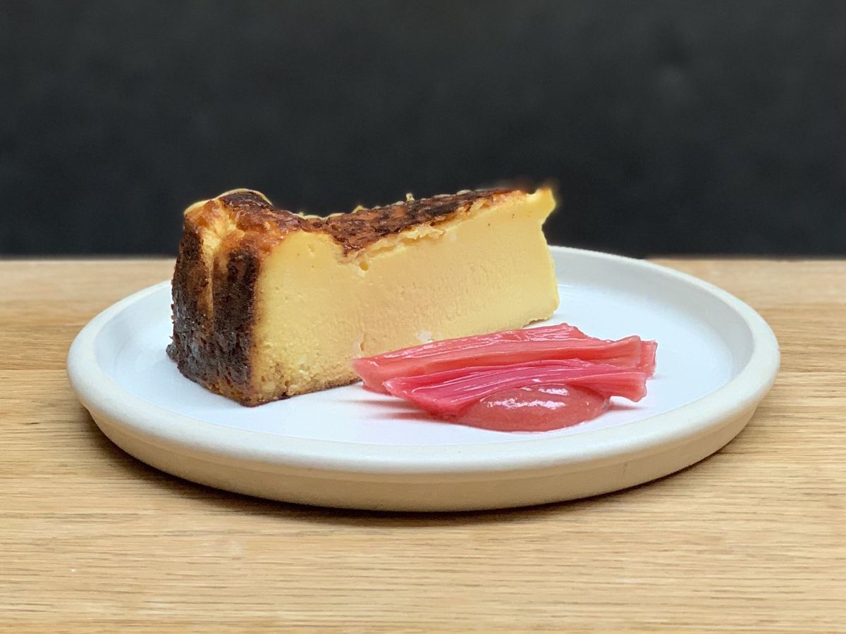 A slice of Basque cheesecake, served on a white deep ceramic plate, with poached rhubarb
