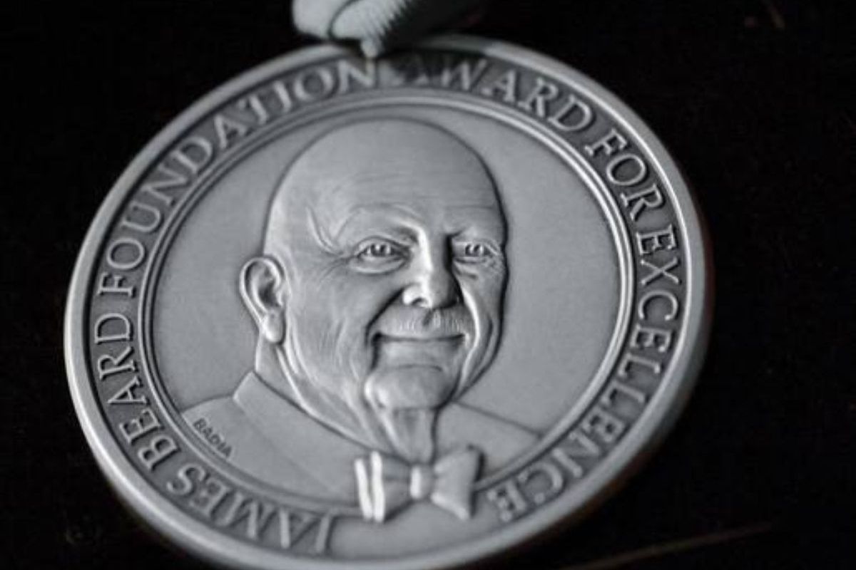 A round silver medal features the face of a bald, older man wearing a bowtie. Words run around the edge, reading James Beard Foundation Award for Excellence.