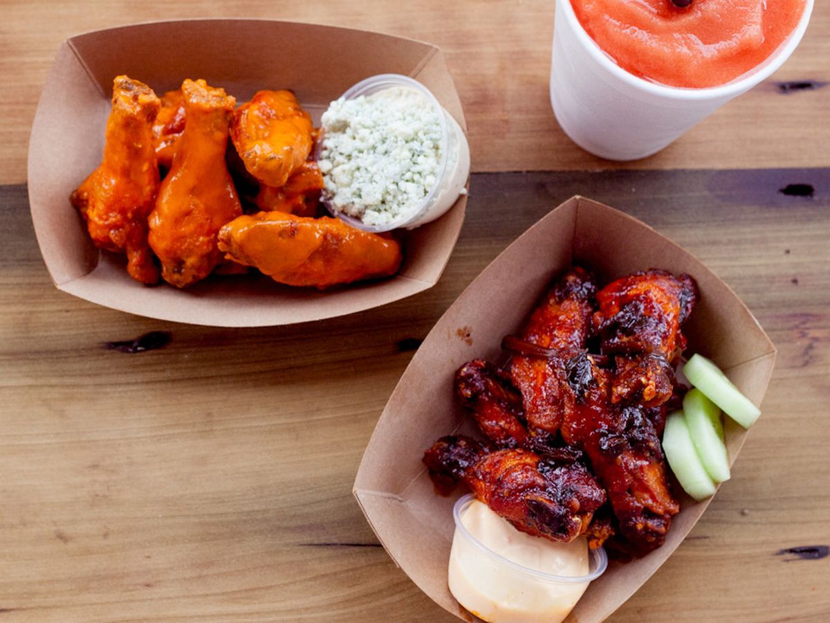 Overhead view of two cardboard trays of wings; one light orange with a side of blue cheese crumbles and one dark red/brown and charred with celery sticks.
