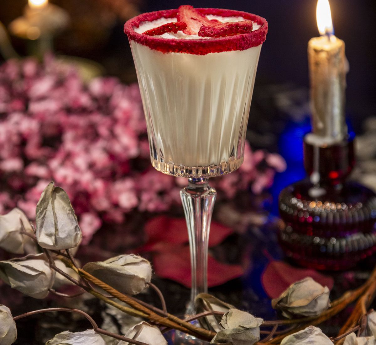 A white cocktail rimmed with red sprinkles, set near roses and candles.