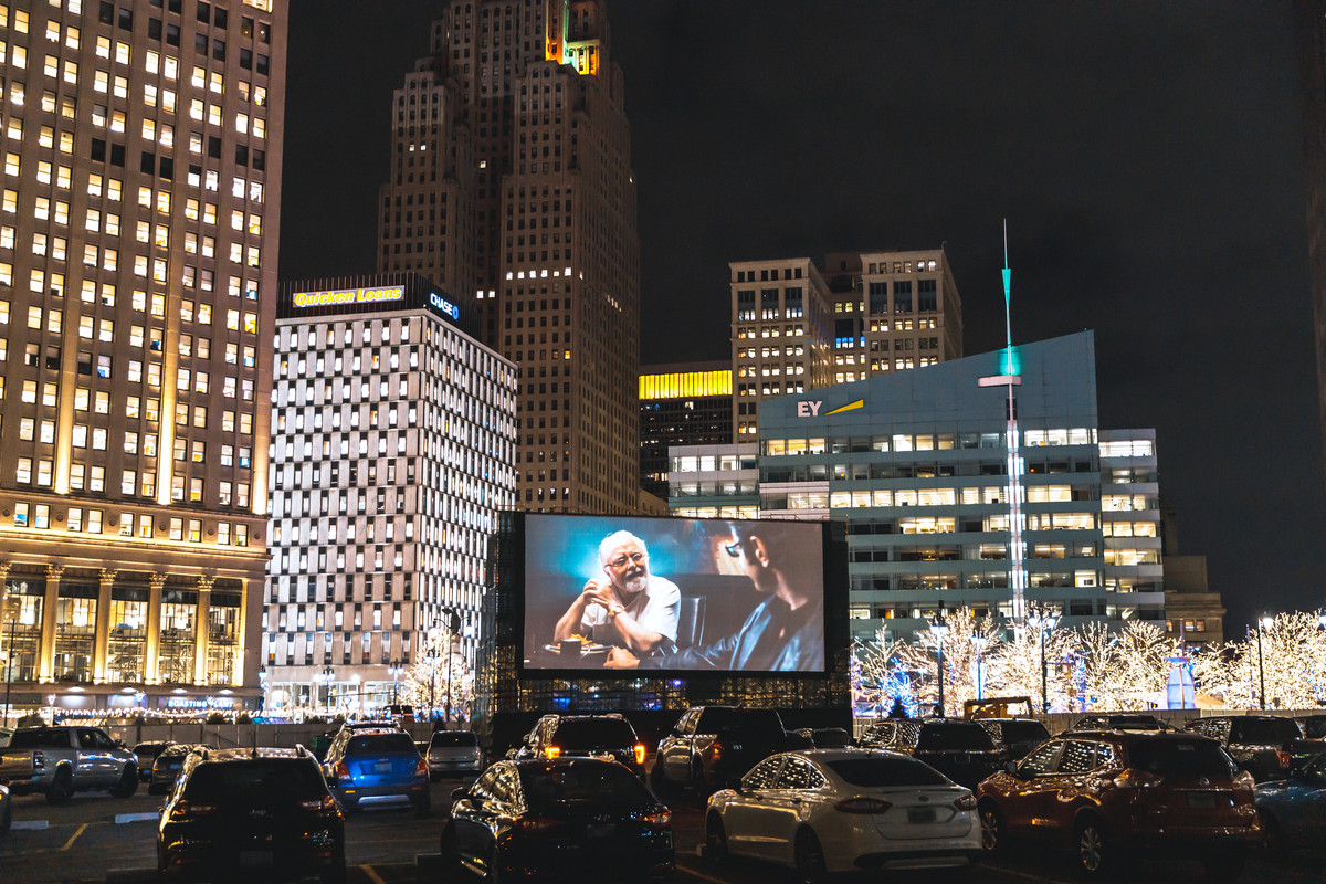 On a clear, dark night against the backdrop of the lit up skyline of downtown Detroit, cars line a parking lot to watch Jurassic Park at the Monroe Street Drive-In. 