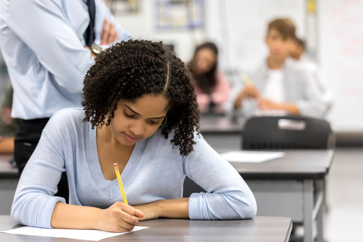 A young girl sit at a black desk in a classroom taking an exam with a yellow number two pencil. A teacher stands behind her monitoring the classroom. Other students are unfocused in the background taking the exam.