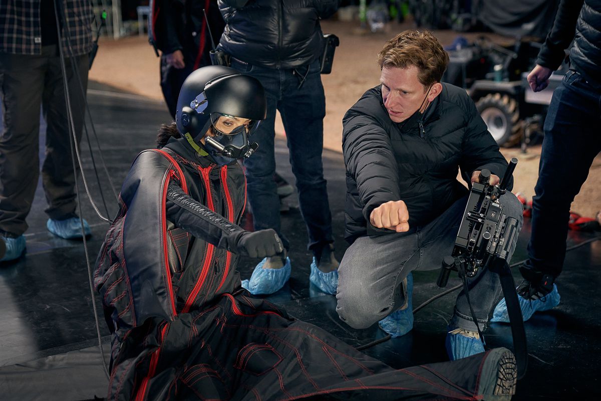 Gal Gadot and Tom Harper point together while filming Heart of Stone. Gadot wears a skydiving suit and a helmet.