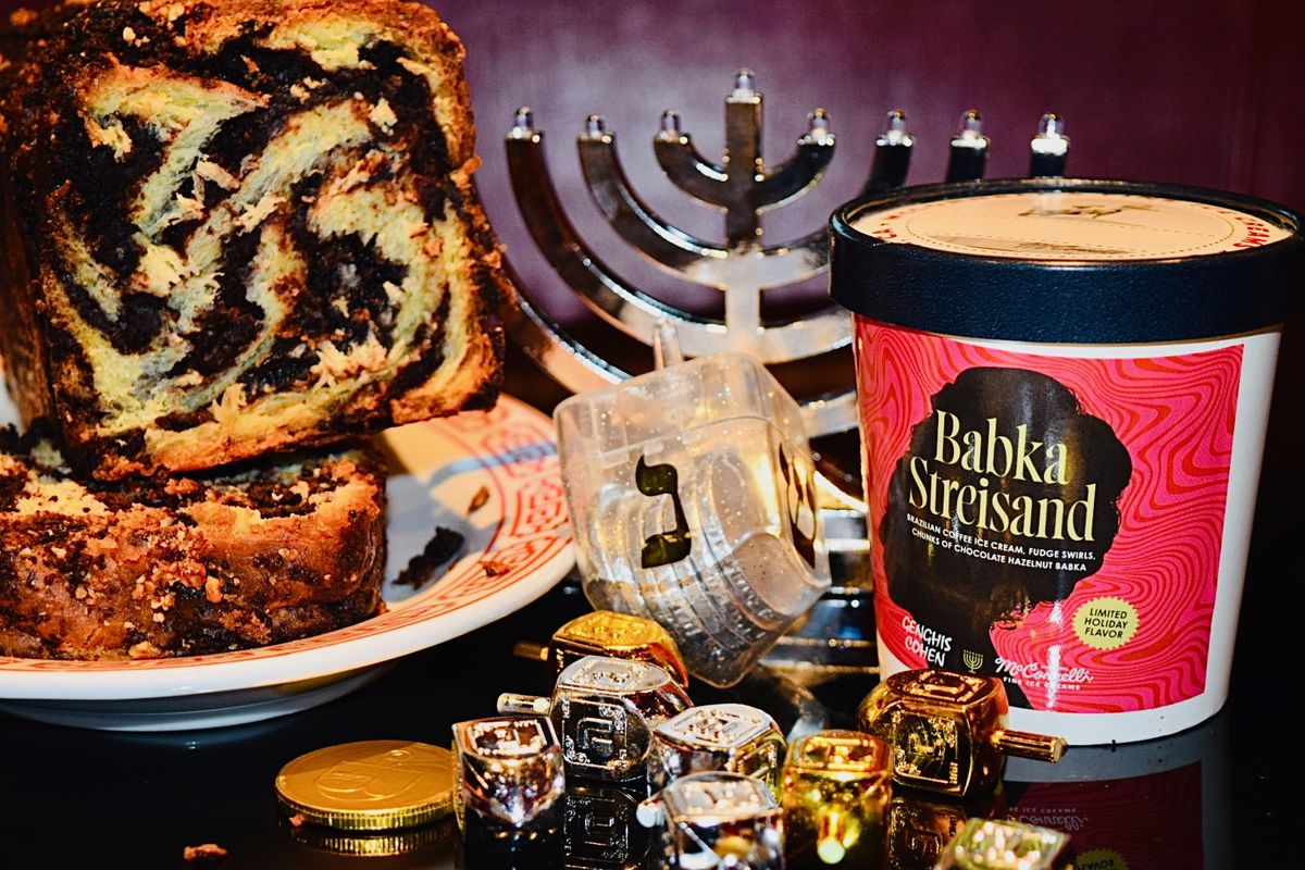 A plate of chocolate babka and a red-printed ice cream pint labeled “Babka Streisand” bookend a table filled with Hanukkah ephemera: a silver menorah, a crystalline dreidel, and a table scattered with mini dreidels and Hanukkah gelt.