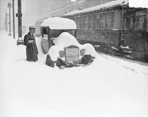 Image of a man standing in knee deep snow next to an automobile with snow piled on the roof, hood and around the wheels on a street in&nbsp;Chicago,&nbsp;Illinois. A streetcar (trolley car) is moving through the snow past the automobile on the right.