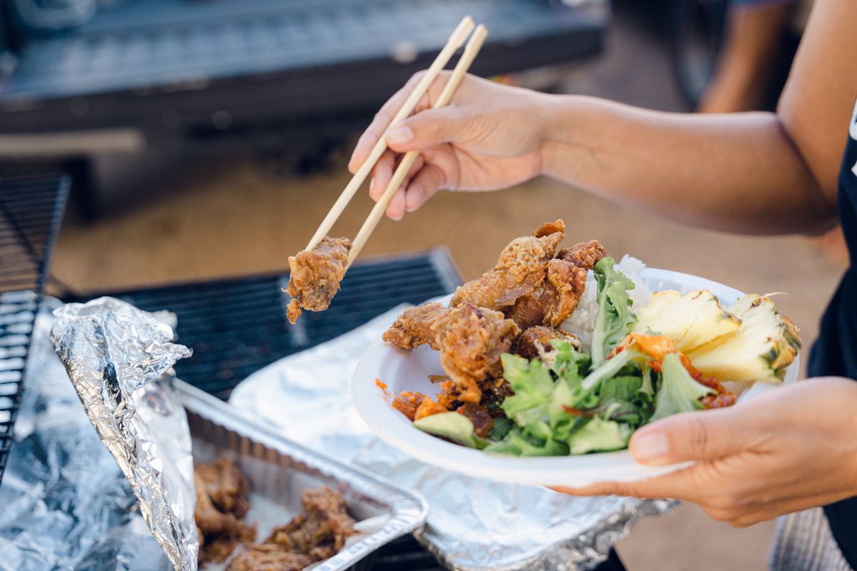 Hand holding pair of chopsticks grabs piece of fried chicken from a foil-lined tin; the other hand holds a disposable plate containing fried chicken, lettuce leaves, and pineapple wedges.