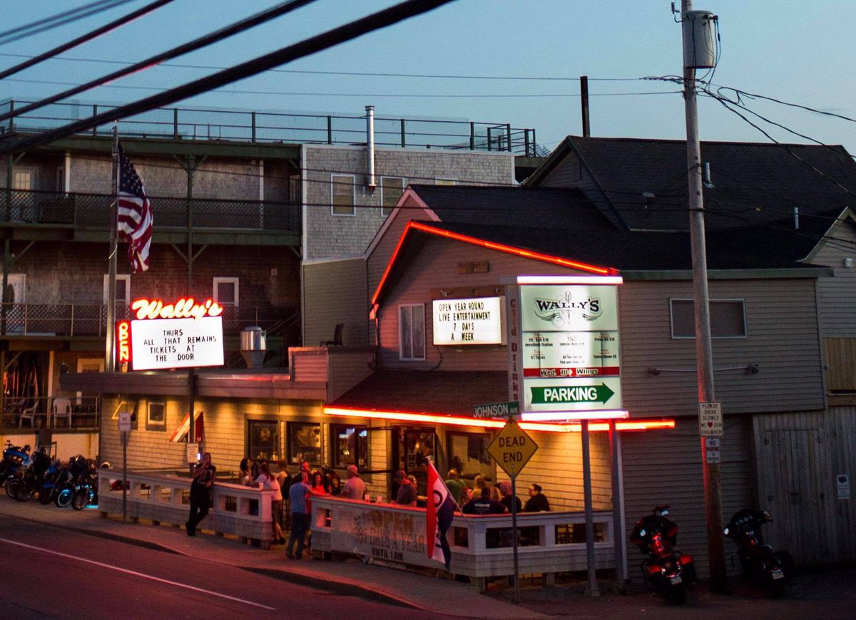 Exterior of a bar at dusk, with patrons on a patio out front. A letterboard sign is topped with neon red signage reading Wally’s in cursive.