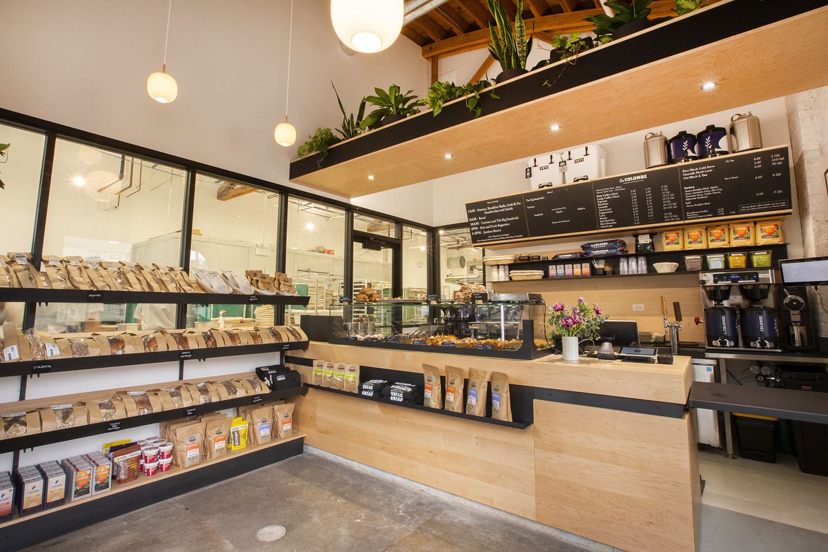 A wooden cafe counter with coffee urns behind, a pastry case on top, and shelves off to the side that hold loaves of bread.
