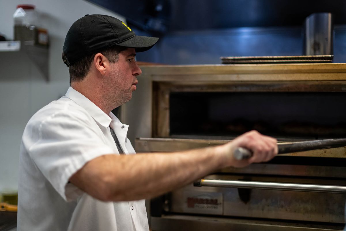 A man in a white shirt and black hat turns a pizza inside of a deck oven at a restaurant.