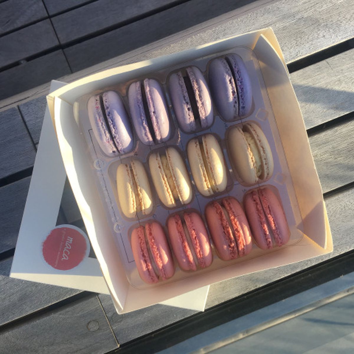 A white box is open to reveal a dozen macarons —&nbsp;four each in pale purple, off white, and pale pink. The box sits in the sun on a picnic table.