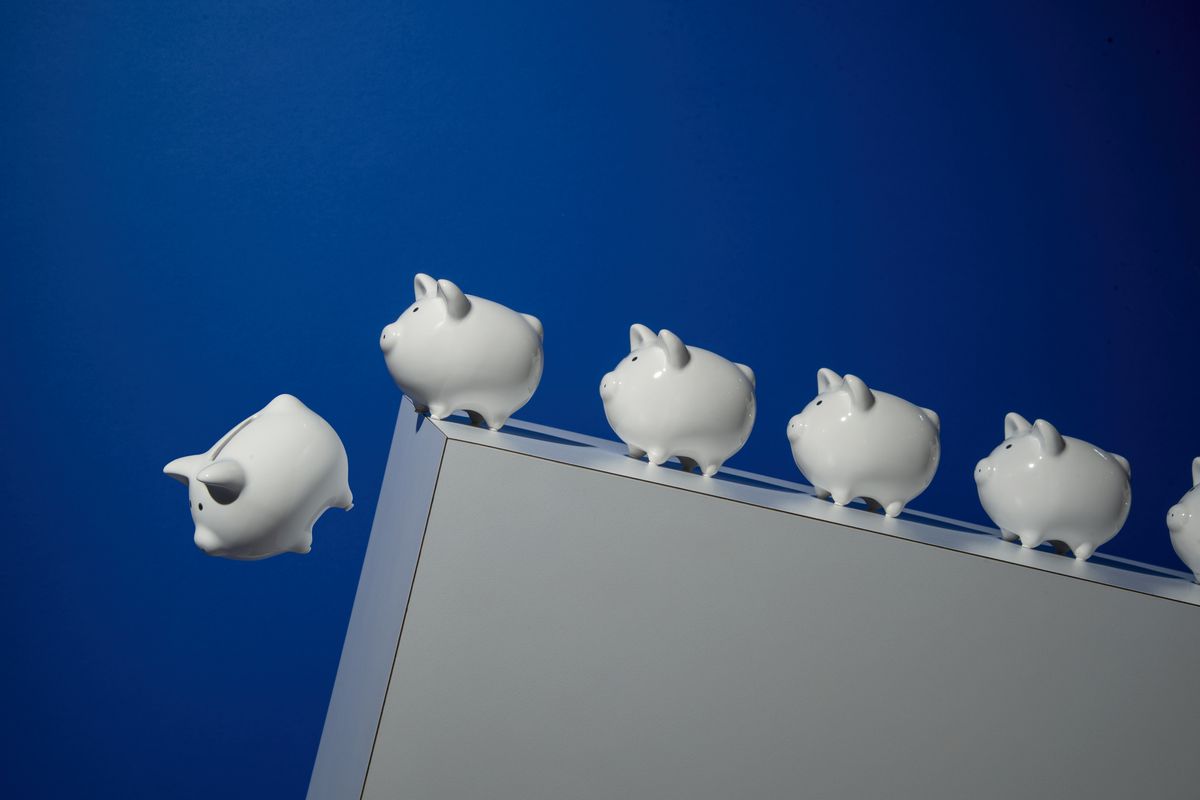 A line of porcelain piggy banks stand at the edge of a rectangular cliff. One has fallen off the cliff, pictured mid-air.