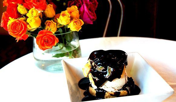 A cream puff topped with chocolate sauce sits on a white plate on a white tablecloth. A vase of colorful flowers sits in the background.